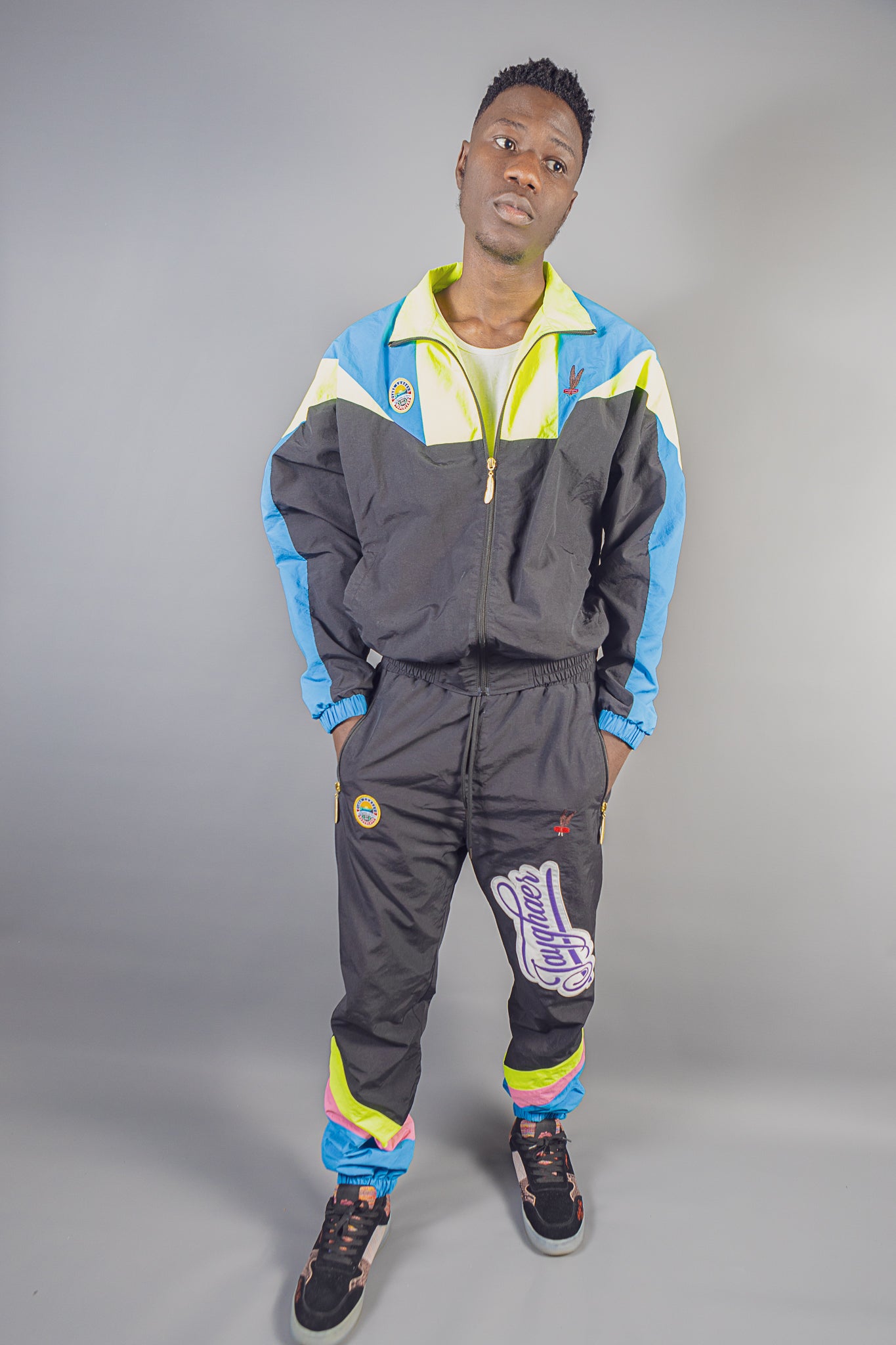 Man wearing JayghaerPierre black tracksuit with blue, yellow, and pink accents against a gray backdrop