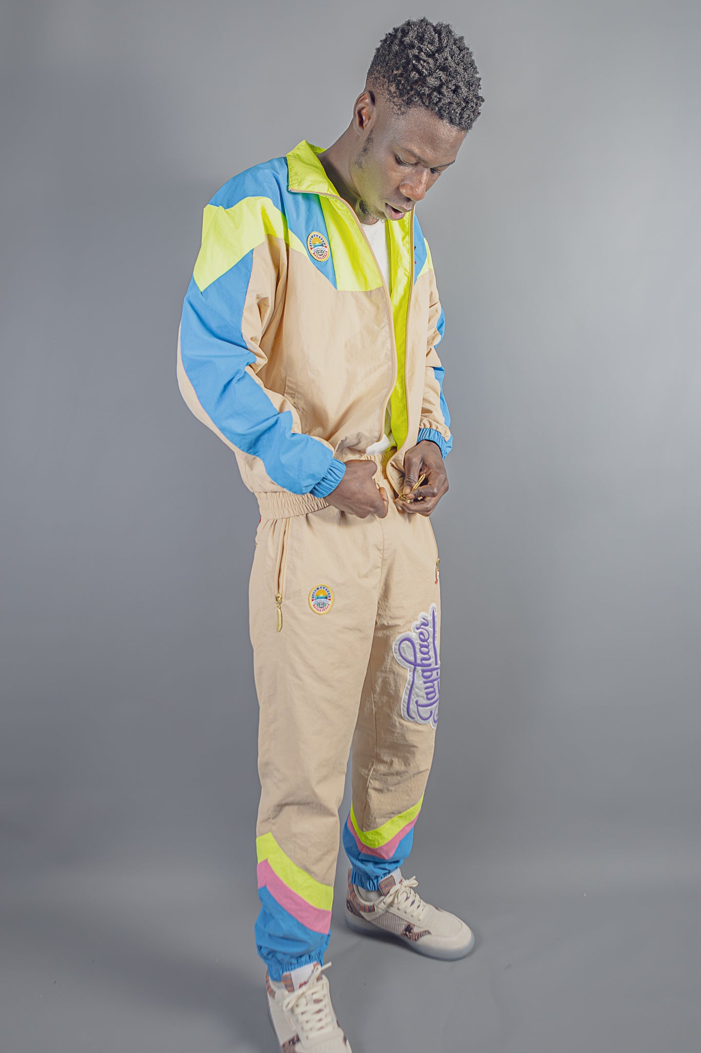 Man wearing beige, blue, neon yellow, and pink Jayghaer Pierre tracksuit with logo patches against gray background