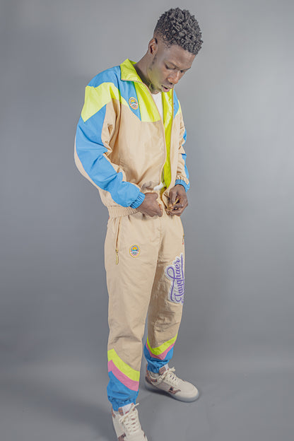 Man wearing beige, blue, neon yellow, and pink Jayghaer Pierre tracksuit with logo patches against gray background