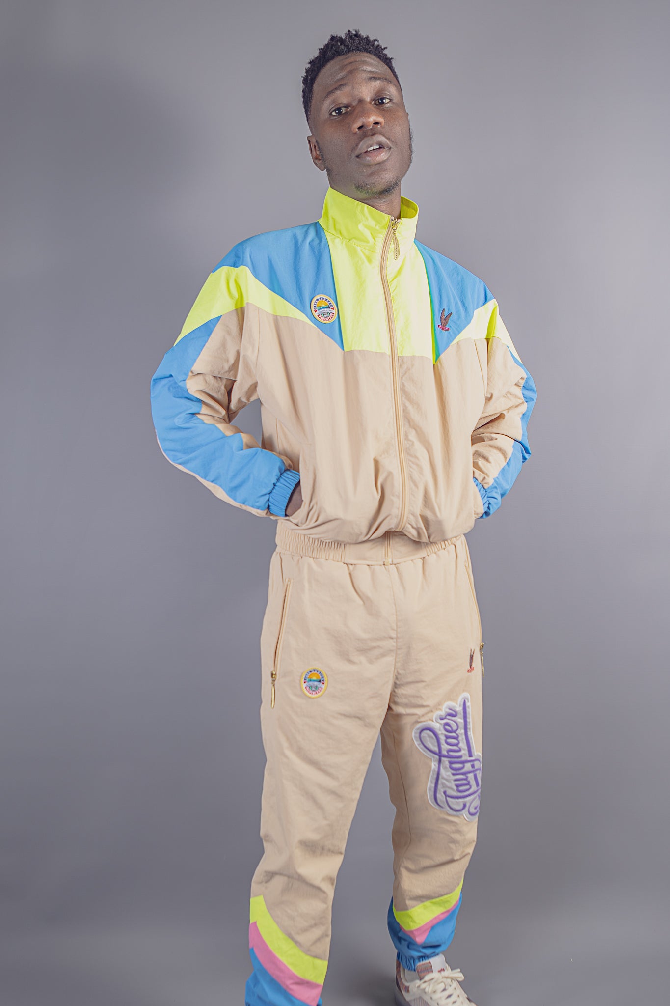 Man wearing beige, blue, and yellow color-blocked tracksuit with patches and zippers on grey backdrop