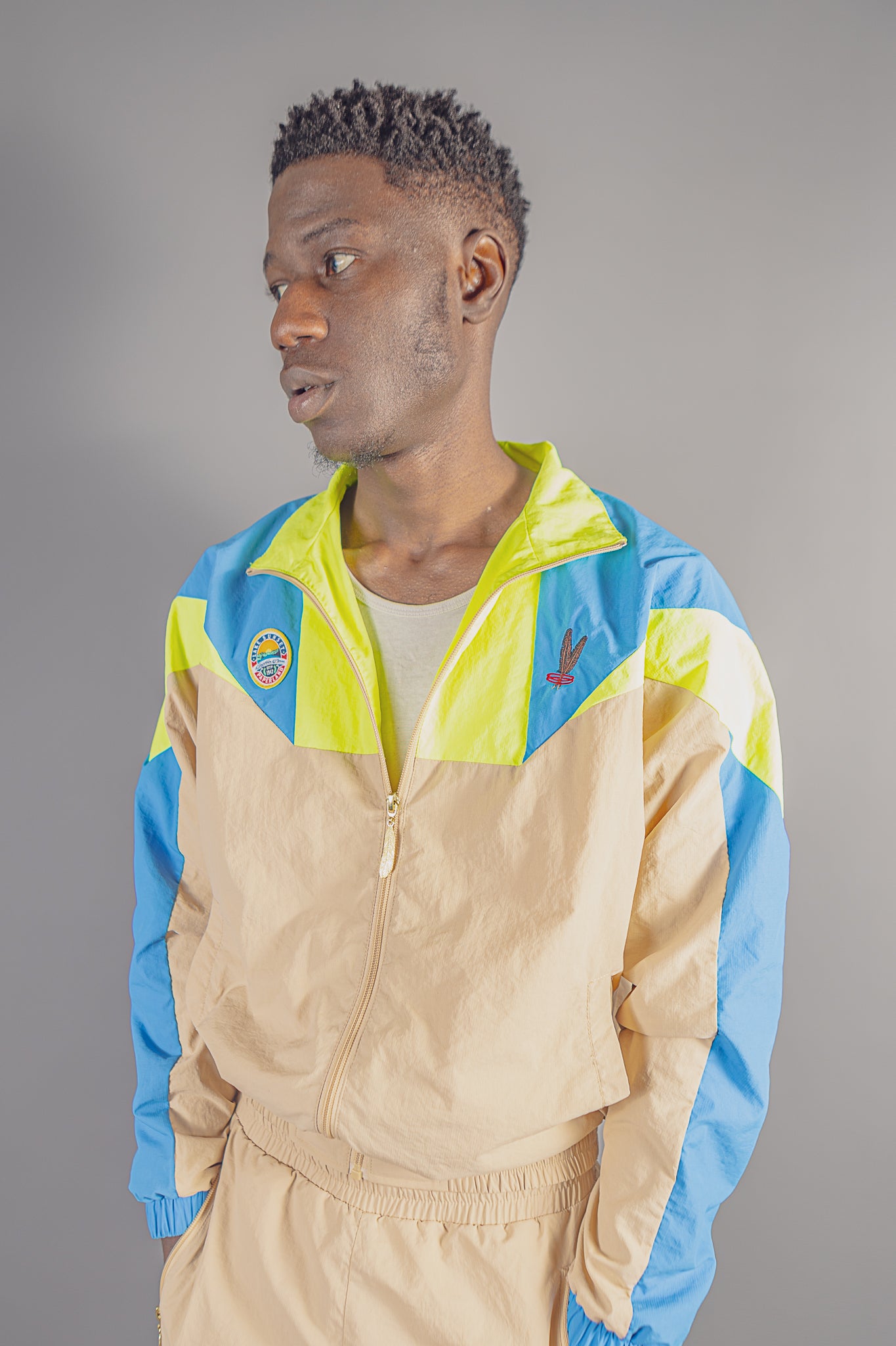 Man wearing beige, blue, and neon yellow zip-up jacket with JayghaerPierre logo on gray background