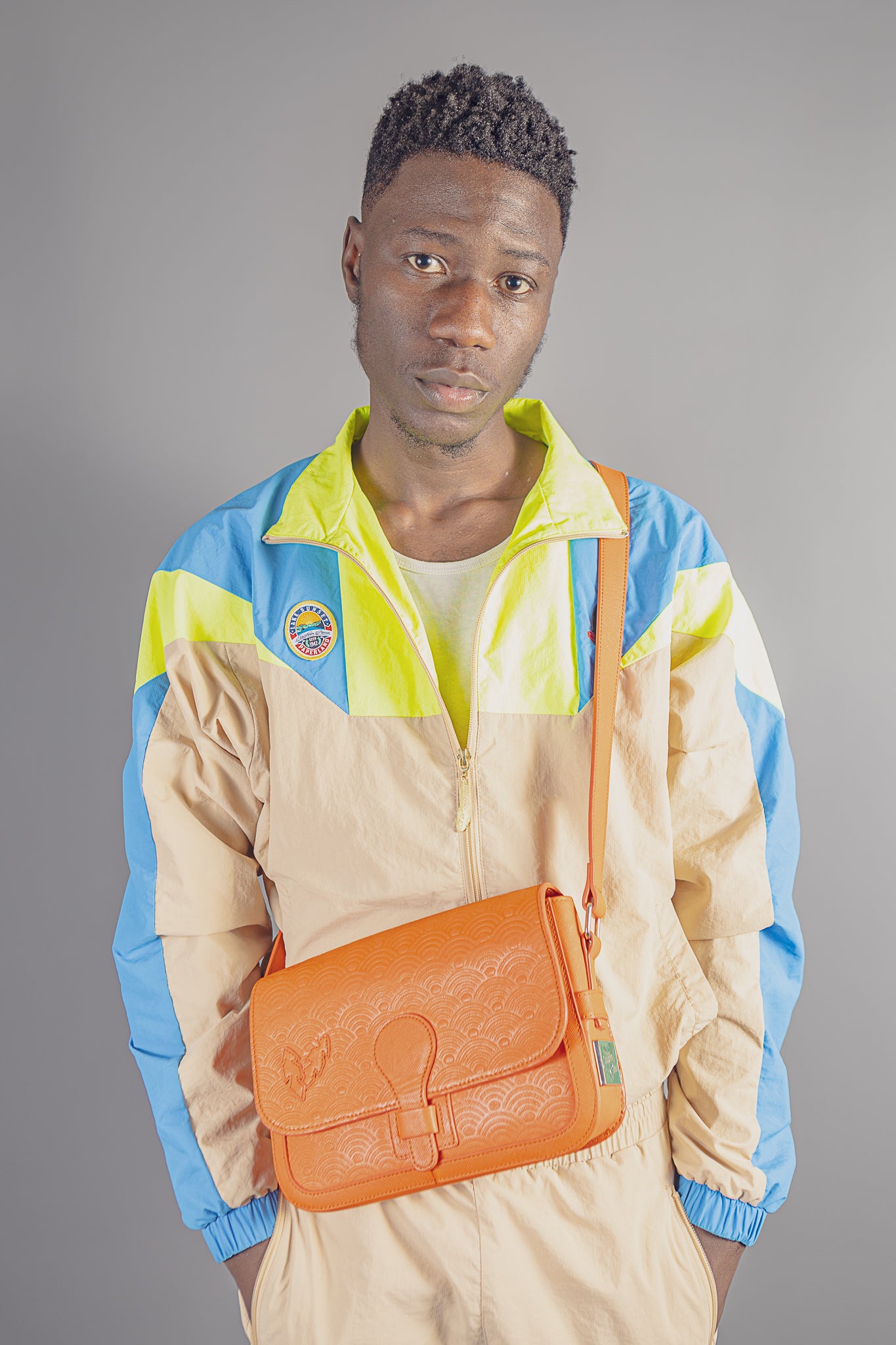 Male model in colorful windbreaker jacket with bright orange embossed leather crossbody bag on gray background