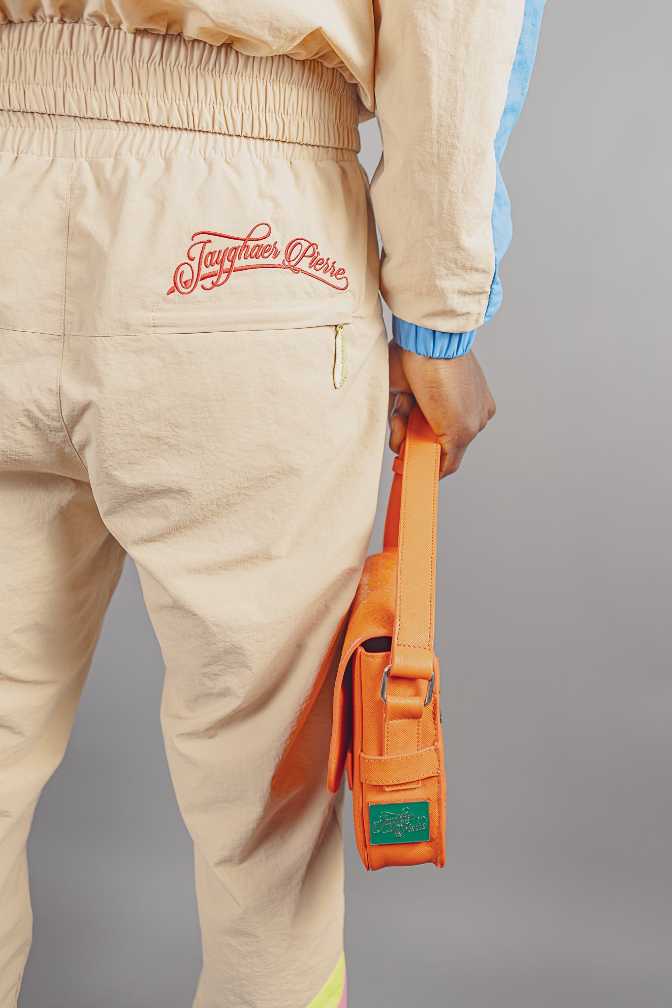 Beige Jayghaer Pierre pants with elastic waistband and red logo embroidery, model holding orange Jayghaer Pierre bag