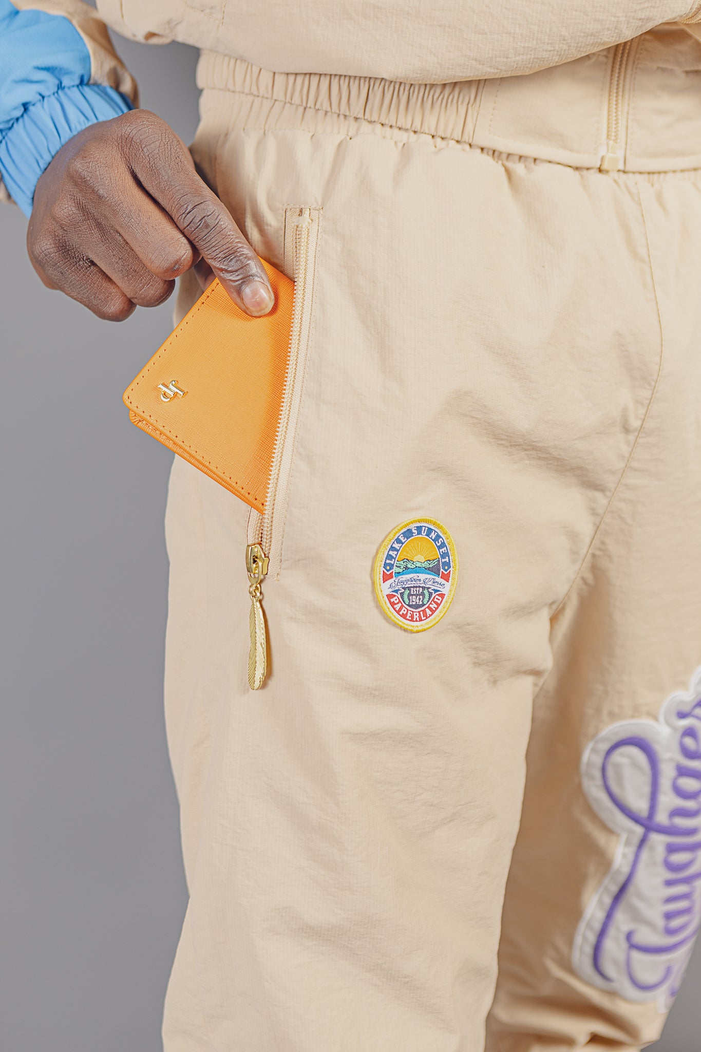 Hand placing orange wallet into beige pants pocket featuring Lake Sunset Paperland patch